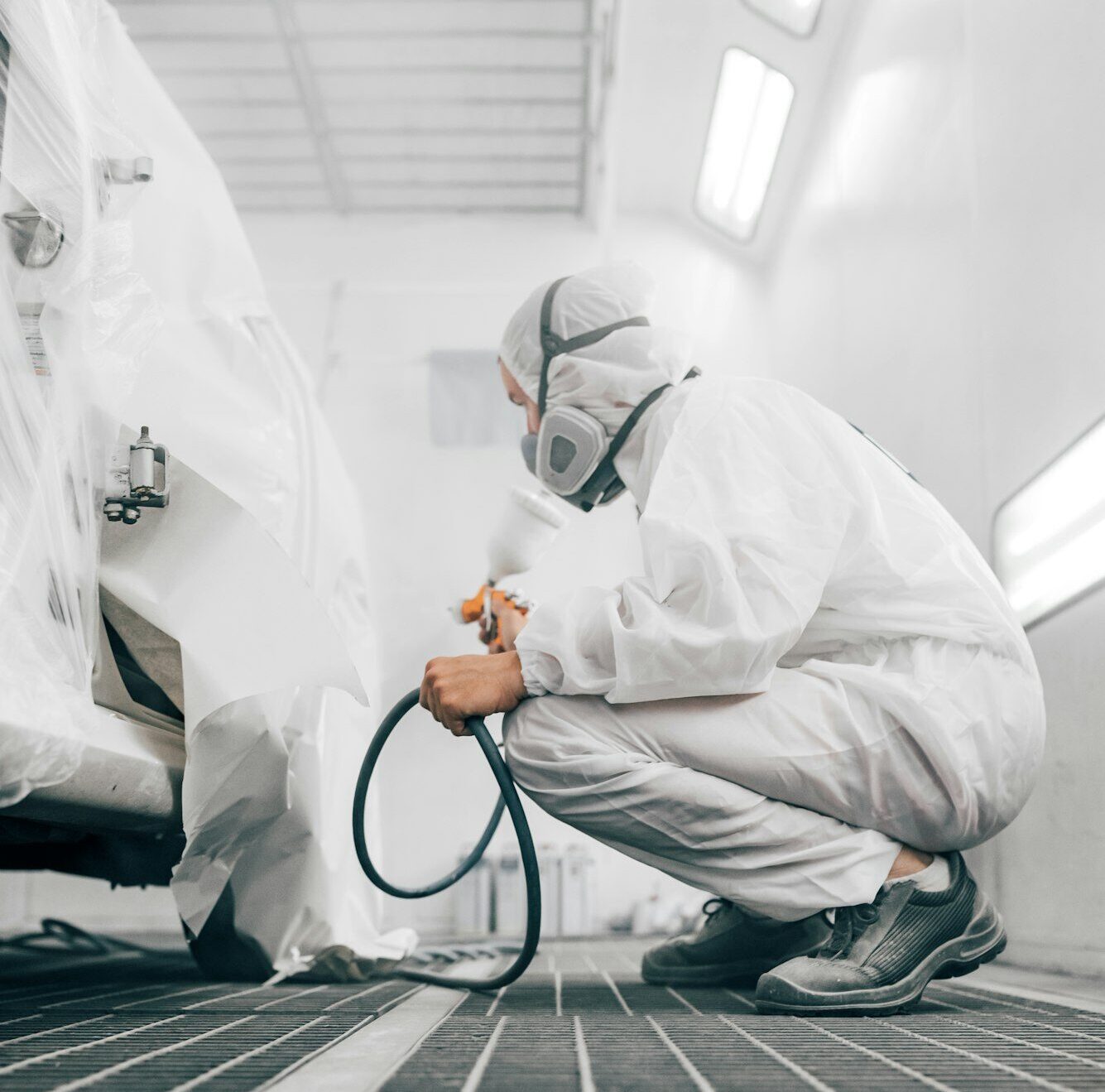 Worker in protective uniform painting car body in paint chamber.